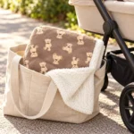 Folded LittleNest Coffee Bear baby blanket in a changing bag beside a stroller